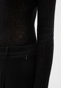 Melna, ar ribojumu, ar garām piedurknēm, ierakta augsti piegulošās melnās biksēs ar jostas cilpu un sānu kabatas detaļu.