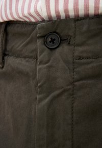 Pantaloni in tessuto khaki con un bottone nero in vita, con texture diagonale e una leggera lucentezza, camicia a strisce parzialmente visibile sopra.