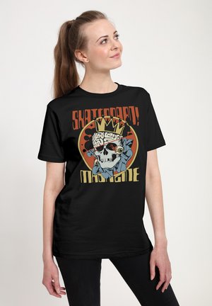 Žena s dlouhými vlasy svázanými dozadu nosí černé tričko s lebkou s korunou a nápisem „Skateboard! Masque“.