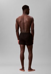 Czarne kąpielówki z elastycznym pasem, wykonane z lekkiego materiału. Model jest nagi i boso, pokazany od tyłu.