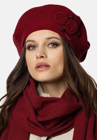 Berretto rosso lavorato a maglia con tre accenti floreali e una sciarpa abbinata. Tessuto testurizzato con una finitura liscia, progettato per adattarsi comodamente.