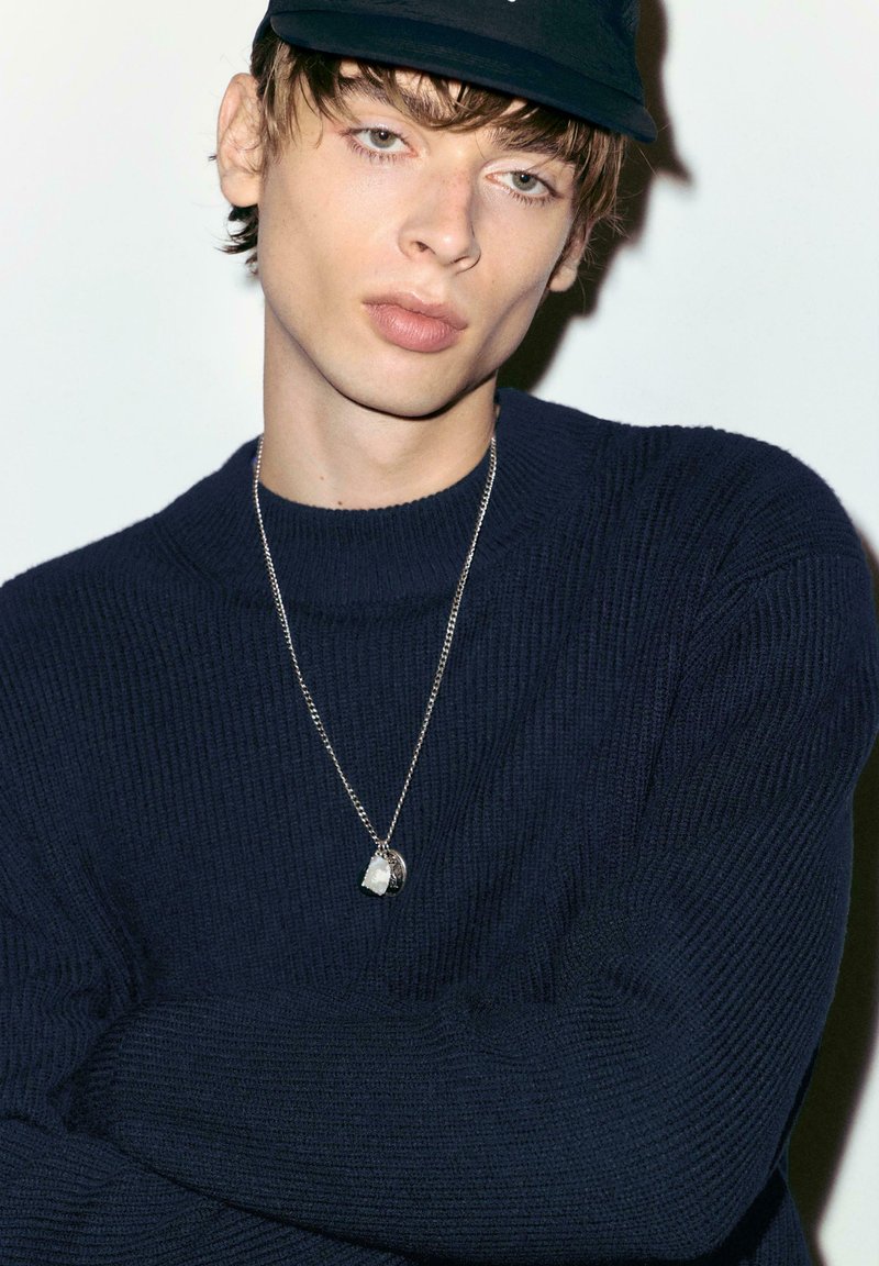 Pull en maille bleu marine avec col montant, collier à chaîne argentée avec pendentif décoratif, et casquette noire sur un fond neutre.