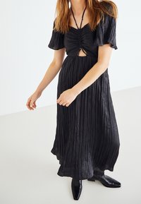 Vestido negro con mangas cortas con volantes, cuerpo fruncido, detalle en forma de corte y tejido texturizado; largo hasta el suelo y combinado con botines negros.