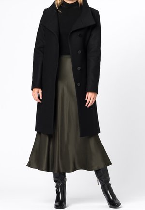 Femme portant un manteau noir boutonné sur un haut noir, une jupe midi en satin vert olive et des bottes en cuir noires mi-mollet, debout devant un fond blanc.