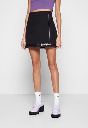 Femme portant une mini-jupe noire avec bordure blanche et logo "Tommy", haut court violet, chaussettes blanches Nike, et mocassins à plateforme lilas.