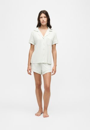 Femme debout pieds nus portant une chemise légère à manches courtes boutonnée et un short assorti sur fond blanc uni.