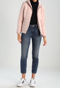 Giacca a vento rosa chiaro con zip frontale e cappuccio, abbinata a jeans blu scuro e sneakers bianche con strisce nere.