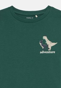 Zelena pamučna majica s okruglim izrezom, na kojoj se nalazi grafika dinosaura u prigušenim bojama i riječ "avantura" u bijeloj boji. Mekana tekstura.