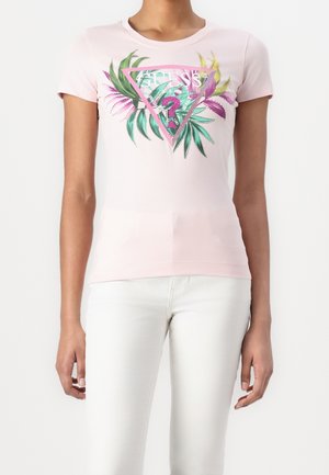 Woman wearing a light pink short-sleeve shirt with a colorful tropical leaf design and Guess logo paired with white pants.
