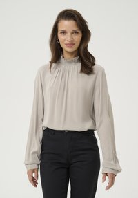 Light gray blouse with a high ruffled collar, long sleeves, and soft pleating. Worn with dark, fitted pants for contrast.