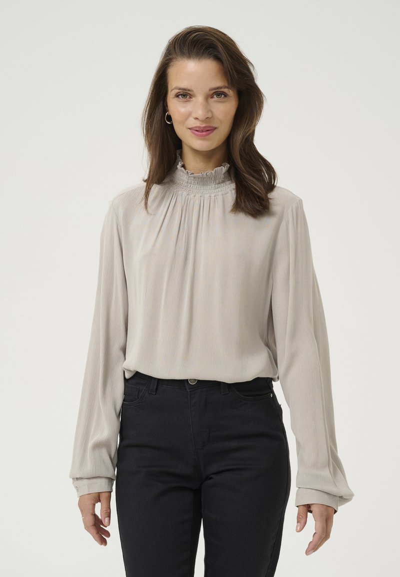 Light gray blouse with a high ruffled collar, long sleeves, and soft pleating. Worn with dark, fitted pants for contrast.