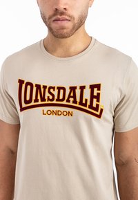 Kreminės medvilninės marškinėlės su apvalia iškirpte, ant priekio puikuojasi didelis bordo ir geltonas "LONSDALE LONDON" logotipas ryškiais raidėmis.