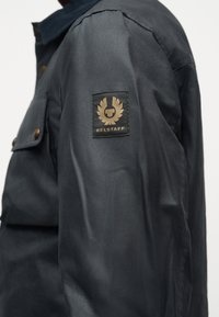 Μαύρο μακρυμάνικο μπουφάν με λογότυπο Belstaff στο πάνω μέρος του αριστερού χεριού και κουμπιά με πατιλέτα από ορείχαλκο στην τσέπη του στήθους.