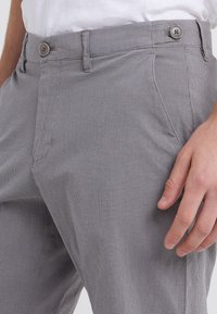 Uomo che indossa pantaloni grigi a quadri con tasche laterali e passanti per cintura, con una camicia bianca infilata, mano appoggiata di lato.