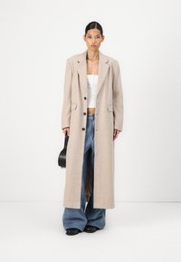 Beige ribbed long coat with notched lapels and black buttons, worn over a white crop top and wide-leg blue jeans, with a black handbag.