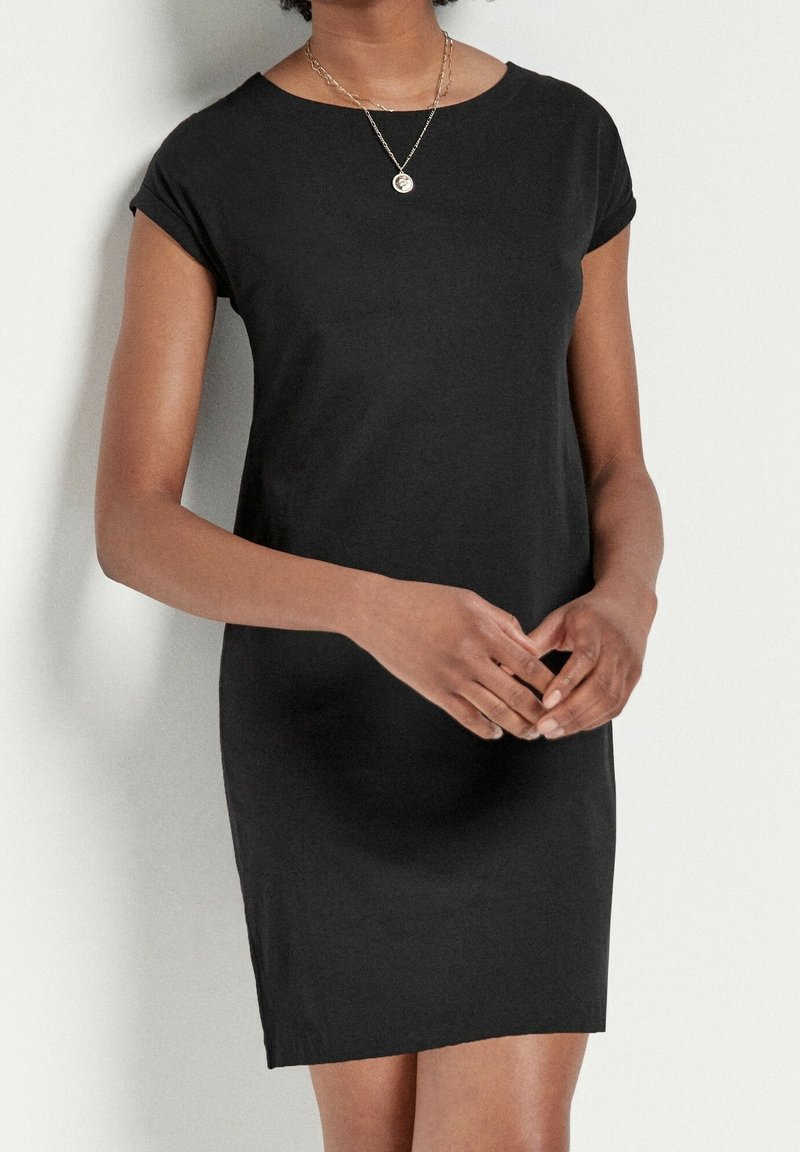 Femme portant une courte robe noire avec des manches retroussées et un collier pendentif rond en or, debout devant un fond clair uni.