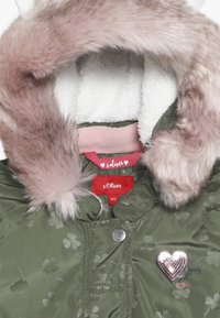 Veste vert olive avec un col en fausse fourrure rose, une doublure en polaire blanche, des motifs de trèfles, et un détail en forme de cœur argenté orné de sequins sur la poitrine.