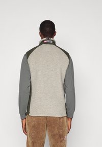 Pessoa vestindo uma jaqueta de fleece cinza e verde oliva com um logotipo vermelho e branco "66 North Iceland" na gola, de costas para a câmara.