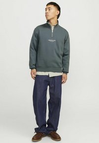Mørkegrøn, sweatshirt med lynlås i halsen lagt over enternet skjorte, parret med løs pasform blå jeans og brune snørebåndssko.