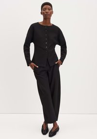 Top lungo a maniche lunghe nero, strutturato, con scollatura rotonda e chiusura a bottoni, abbinato a pantaloni neri a copertura ampia e ballerine nere con borchie.