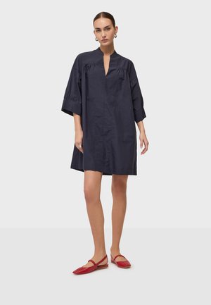 Kobieta w luźnej granatowej sukience sięgającej do kolan z rękawami 3/4 i wycięciem na dekolcie, w połączeniu z czerwonymi balerinami.