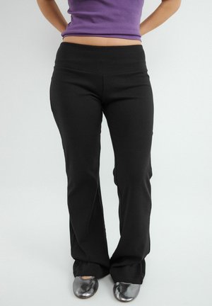 Persoon die zwarte broek met wijde pijpen, zilveren platte schoenen en een paarse cropped top draagt, staand met de handen achter de rug.