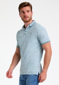 Gaiši zils polo krekls ar rakstainu tekstūru, īsām piedurknēm un apkakli. Aprīkots ar pogām un kontrastējošām svītrām apkaklē.