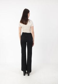 Zwarte flared jeans met een hoge taille, gecombineerd met een aansluitend lichtbeige top. Het model staat van achteren gepositioneerd, zodat de pasvorm goed zichtbaar is.