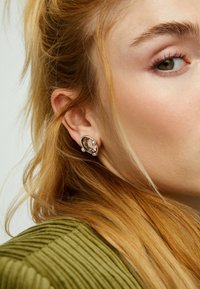 Pendientes de plata con una piedra oval translúcida rodeada por un bisel liso, apoyados contra la oreja del modelo. Tela verde cálida y texturizada visible.