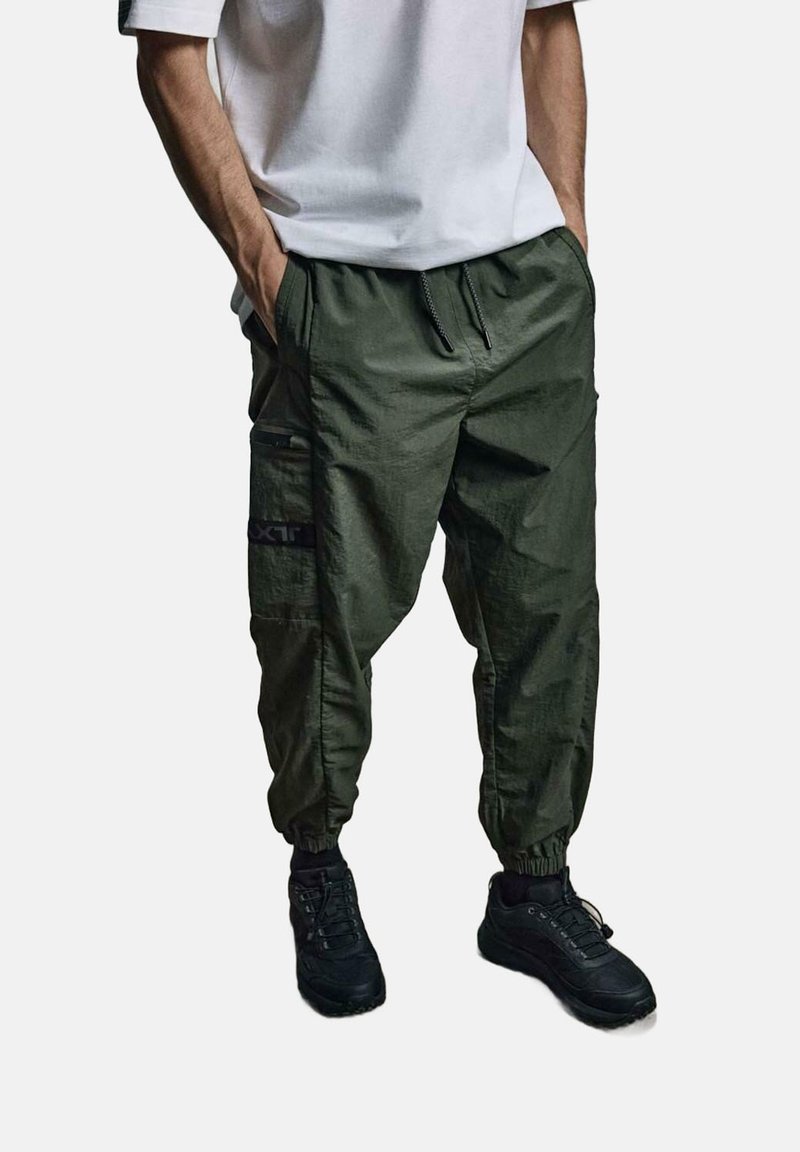 Khaki spodnie cargo z elastycznymi mankietami, wieloma kieszeniami i sznurkiem w talii. Noszone z czarnymi trampkami i białym t-shirtem.