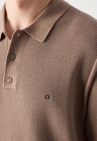 Gros plan d'une personne portant un polo en maille beige texturée avec trois boutons et un petit logo noir "ck" sur la poitrine.