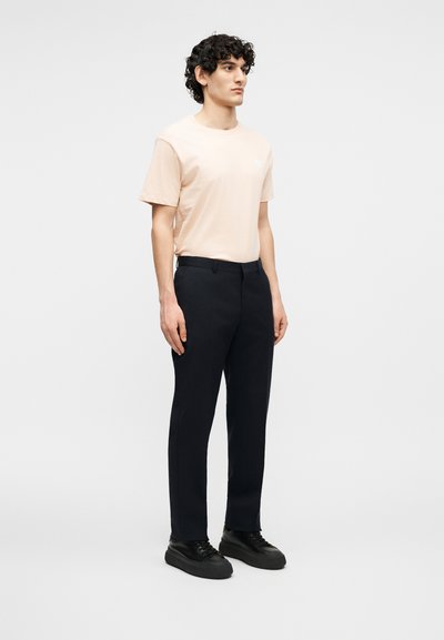 Νέος άνδρας όρθιος, φοράει μπλουζάκι κοντομάνικο σε ανοιχτό μπεζ χρώμα, μαύρο παντελόνι ραμμένο, και μαύρα αθλητικά παπούτσια, σε απλό φωτεινό φόντο.