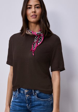 Junge Frau, die ein braunes Kurzarmshirt, blaue Jeans und einen pink-schwarzen, tierfellmusternden Schal um den Hals trägt, vor einfachem Hintergrund stehend.