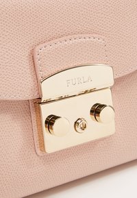 Světle růžová kožená kabelka s texturovaným povrchem, vybavená zlatým kovovým uzávěrem s kruhovými akcenty a vyrytým nápisem "Furla".
