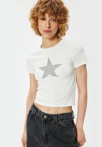 Witte cropped t-shirt met een zilveren glitterster ontwerp. Het model is gecombineerd met zwarte denim jeans, wat een casual uitstraling benadrukt.