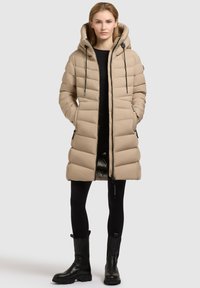 Beige pufferjack met een capuchon, met een gequilt ontwerp, voorzijde met rits, trekkoordsluiting en zijzakken, gecombineerd met zwarte leggings en laarzen.