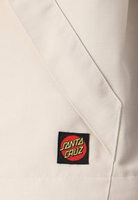 Beige fabric with a subtle textured pattern and black "Santa Cruz" patch featuring red and yellow lettering. Overlapping seam detail visible.