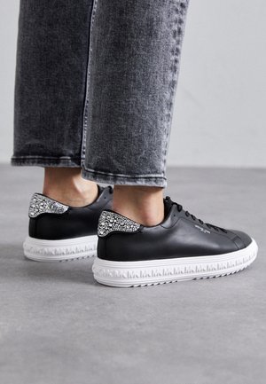 Person iført sorte læder sneakers med hvide såler, prydet med "MK" logo og rhinsten detaljer på hælen, kombineret med grå jeans.
