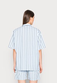 Camisa de manga curta com listas azuis e brancas e um corte descontraído, apresentando um colarinho clássico e riscas escuras em contraste. Usada com calções a condizer.