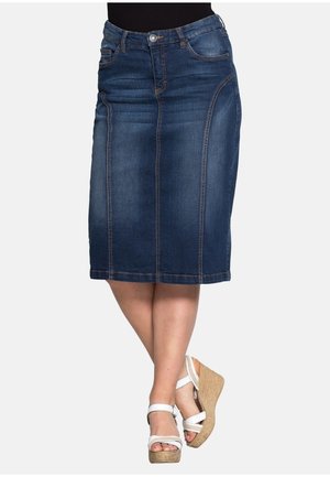 Gonna di jeans blu scuro al ginocchio con dettaglio di cucitura frontale, indossata con sandali bianchi con zeppa su gambe incrociate.