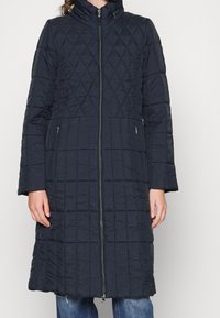 Abrigo largo acolchado de color azul marino con cuello alto, que cuenta con una cremallera frontal y bolsillos laterales, mostrando un patrón de diamantes. Tejido suave e insulado.
