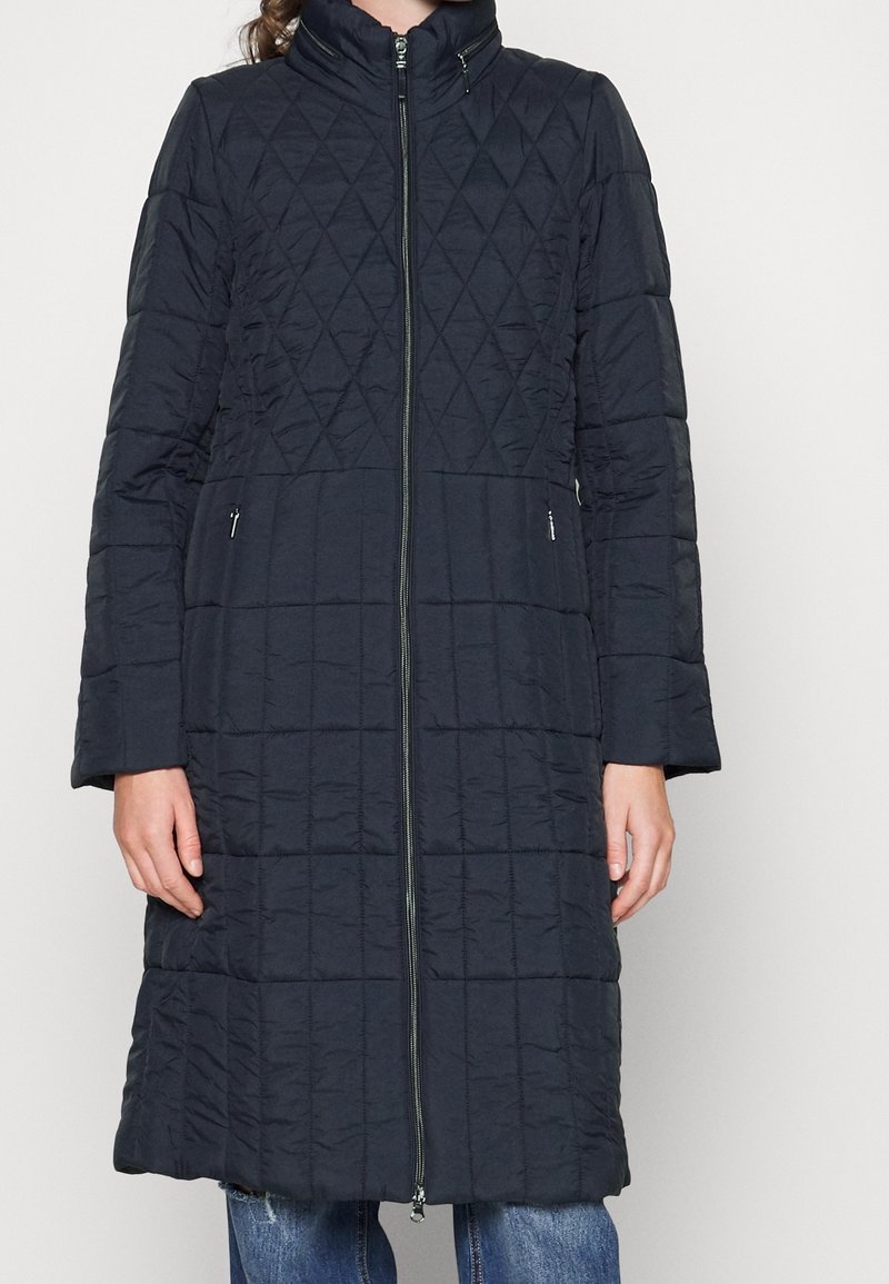 Abrigo largo acolchado de color azul marino con cuello alto, que cuenta con una cremallera frontal y bolsillos laterales, mostrando un patrón de diamantes. Tejido suave e insulado.