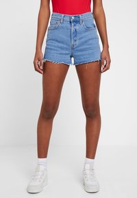 Hellblaue Jeansshorts mit hohem Bund und fransigem Saum, ausgestattet mit vorderen Taschen und einem Knopfverschluss. Kombiniert mit einem roten Oberteil und weißen Sneakers.