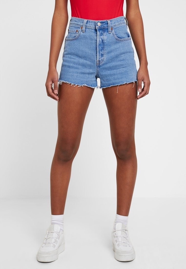 Hellblaue Jeansshorts mit hohem Bund und fransigem Saum, ausgestattet mit vorderen Taschen und einem Knopfverschluss. Kombiniert mit einem roten Oberteil und weißen Sneakers.