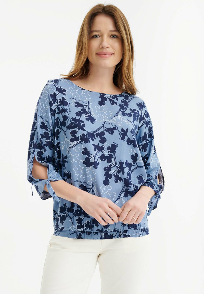 Blusa azul claro con estampado floral en navy, escote barco y mangas abiertas atadas en los codos. Tejido suave y ligero con dobladillo fruncido.