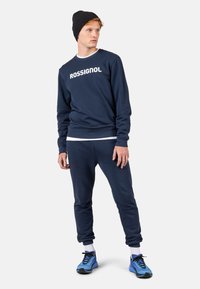 Felpa navy con logo "ROSSIGNOL", abbinata a pantaloni jogger coordinati. Caratterizzata da maniche lunghe, polsini a coste e scarpe da atletica blu.