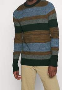 Pull en maille avec des rayures horizontales bleu, marron, gris et vert. Col rond et poignets côtelés. Le tissu présente une texture apparente.