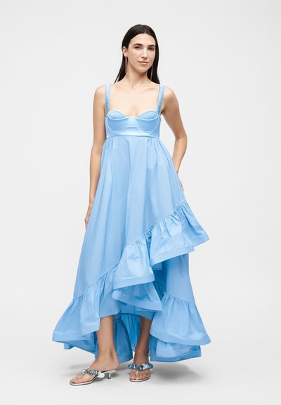 Γυναίκα που φοράει ανοιχτό γαλάζιο ασύμμετρο φόρεμα με βολάν, κορσέ τοπ και ασημένια σανδάλια, στέκεται μπροστά σε λευκό φόντο.