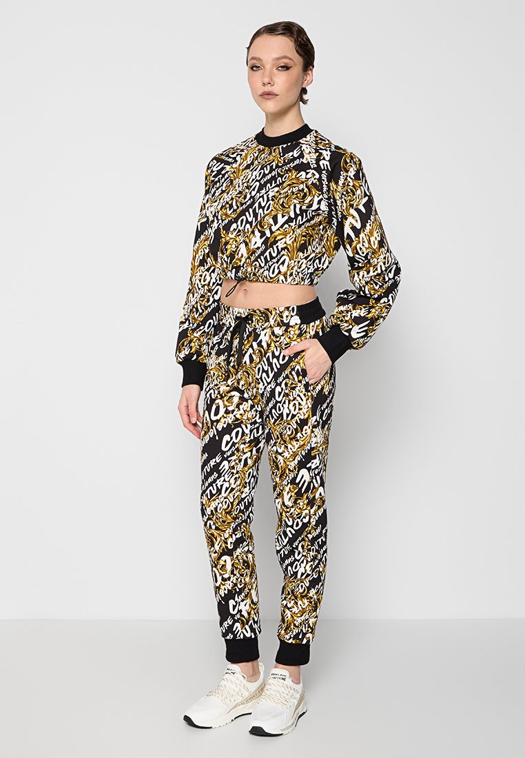 Frau steht da und trägt einen schwarz-weißen und gold gemusterten Cropped-Sweatshirt und passende Jogginghosen mit weißen Sneakers.