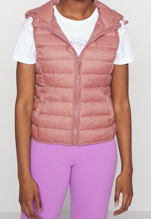 Femme portant une veste matelassée à capuche rose poussiéreux sur un t-shirt blanc et des leggings violet vif, debout devant un fond uni.